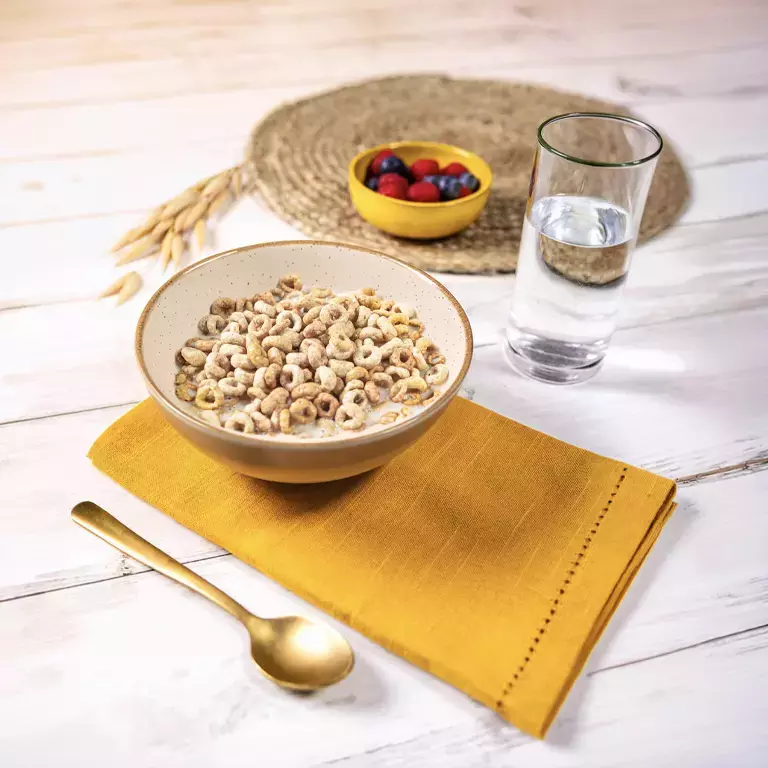 Bol de céréales Cheerios au lait avec verre d’eau, fruits rouges et cuillère sur une table en bois.