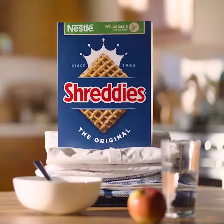 A Box of Shreddies Original on a breakfast table with a bowl, spoon and apple