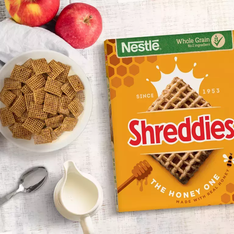 A bowl of Honey Shreddies cereal next to a pack on a table with two apples.