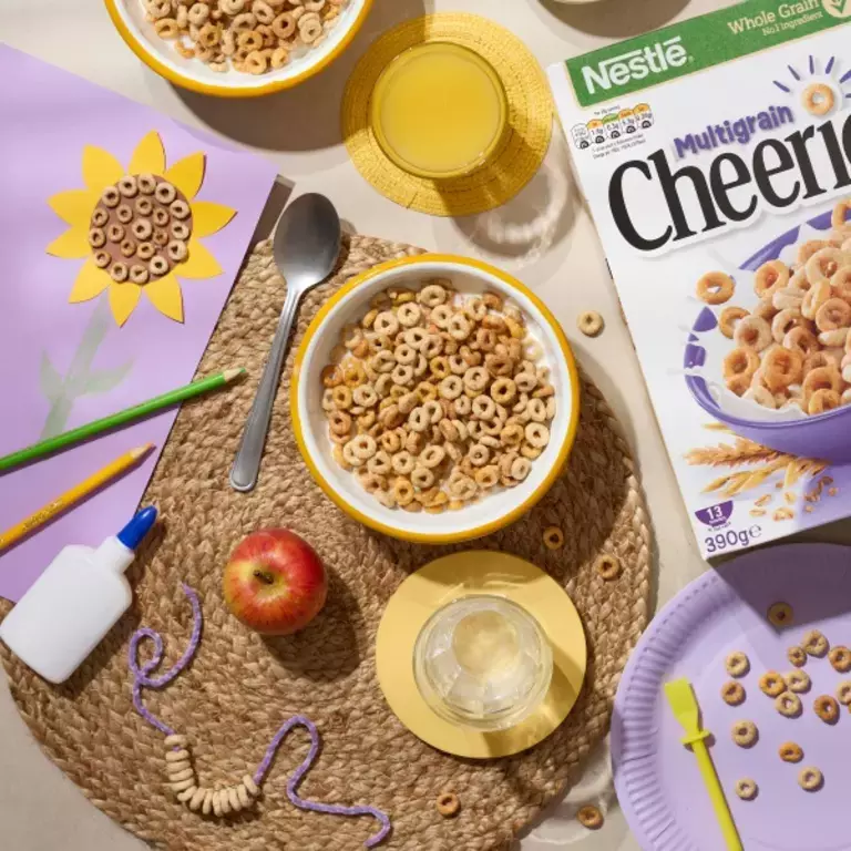 Cheerios with milk in a bowl, with orange juice and an apple. School art project with sunflower drawing and glue alongside.