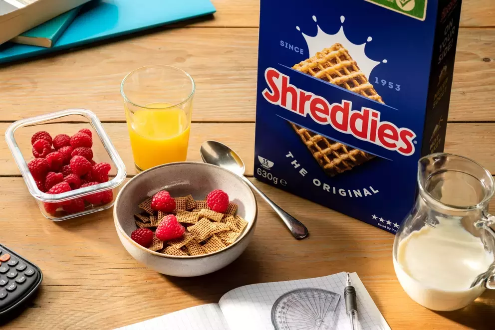 A bowl of Shreddies cereal next to a box of Shreddies with a spoon, a glass of orange juice and a container of raspberries.